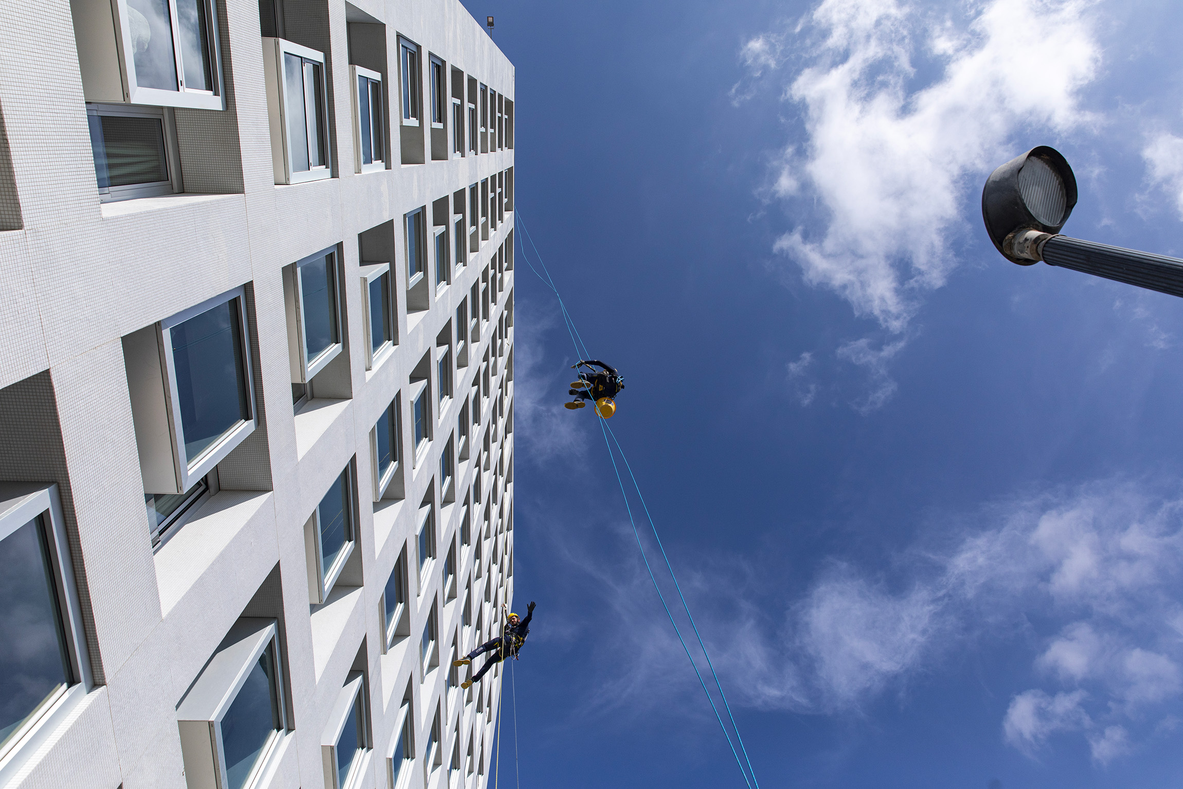 Operatori su fune si calano lungo la facciata bianca di un edificio alto, con il cielo azzurro sullo sfondo