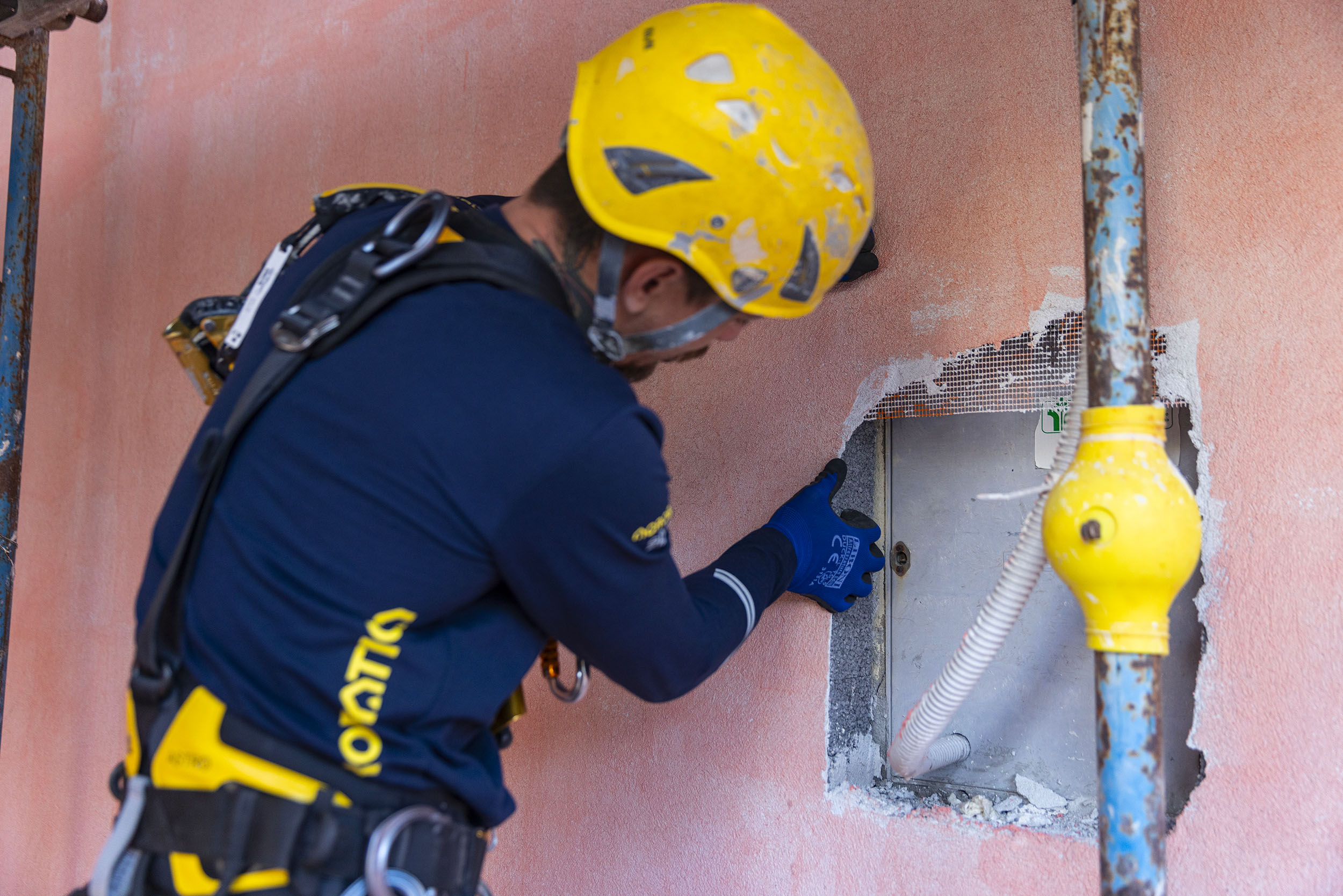 Operatore su fune con imbracatura e casco giallo controlla un quadro tecnico a parete