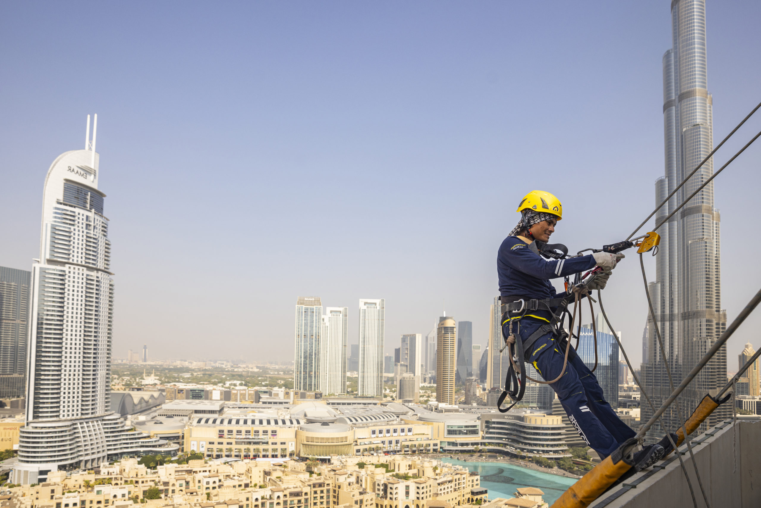 Operatore con casco giallo e imbracatura al lavoro in quota, con lo skyline di Dubai sullo sfondo